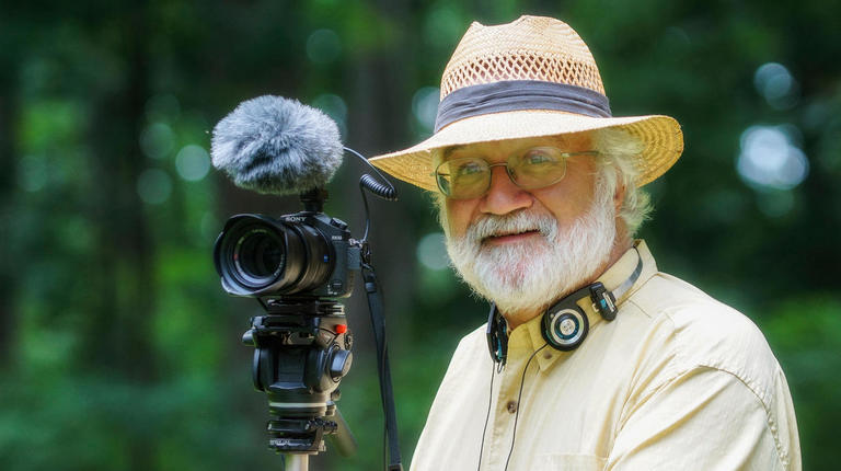 Picture of nature photographer Bob Krist, wearing a straw hat and yellow shirt, standing with his camera in front of a forest during daytime.